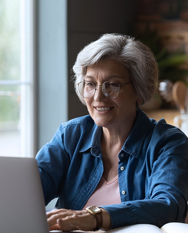 Smiling mature woman with gray hair wearing glasses and denim shirt at laptop near window.