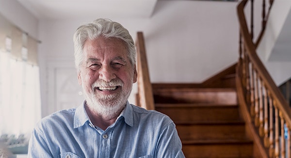 Smiling senior man with white hair in light blue shirt, standing in home interior with wooden staircase.