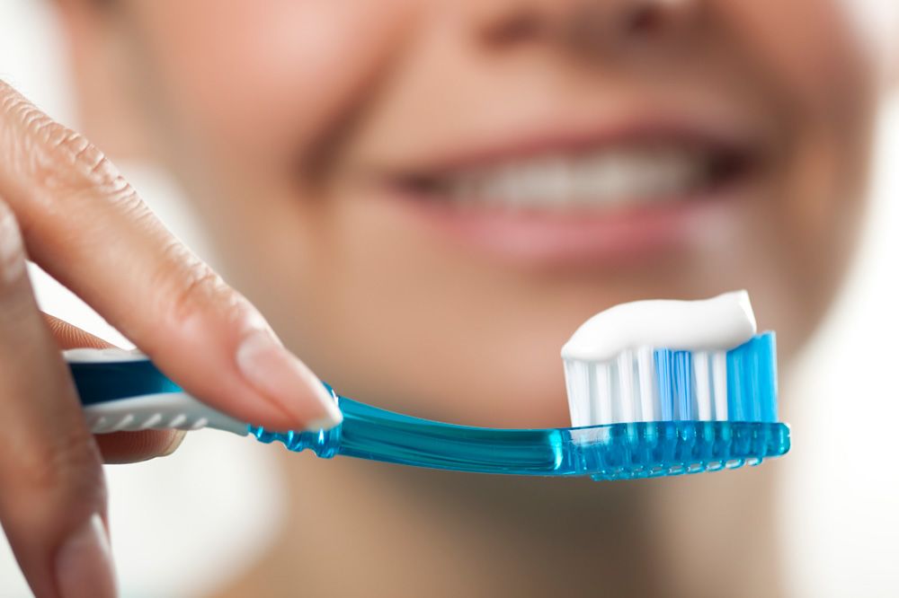 Person holding blue toothbrush with white toothpaste, smiling in background.