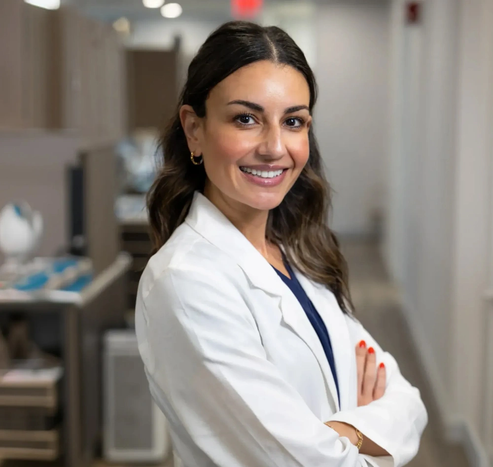 Professional woman in white lab coat smiling with arms crossed in medical office hallway.