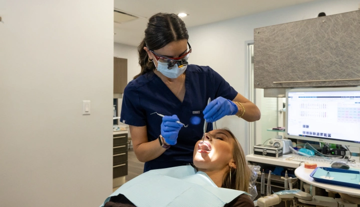 Dental hygienist in blue scrubs and mask examining patient's teeth in modern dental office.