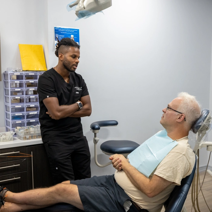 Dental professional in black scrubs consults with older male patient seated in dental chair.